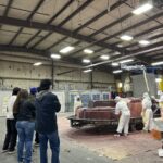 A group of people observe workers in protective gear assembling or coating large molds inside an industrial warehouse.