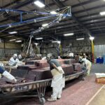 Workers in protective gear assemble or finish large, red, rectangular components inside an industrial warehouse with overhead equipment and bright lighting.