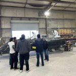 A group of people stand near a boat hull inside a workshop while two individuals in protective suits work on the boat.