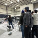 A group of people listens to a woman speaking inside a large workshop with boats and equipment visible in the background.