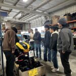 Six people stand in a workshop, gathered around a yellow and black machine placed on a wooden crate, with shelves and tools visible in the background.
