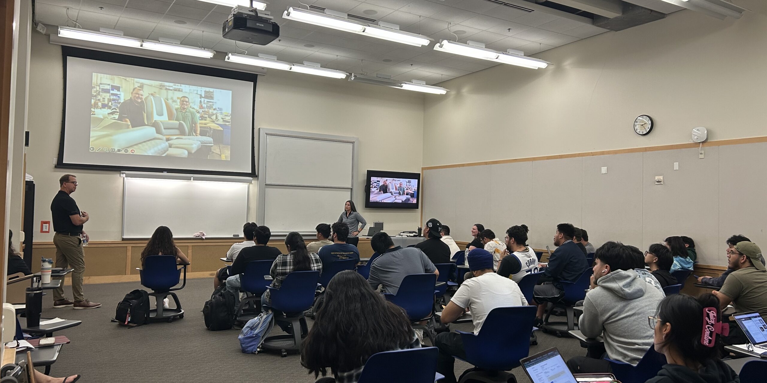 A classroom with students seated at desks facing a lecturer and presentation screen; some students use laptops while listening.