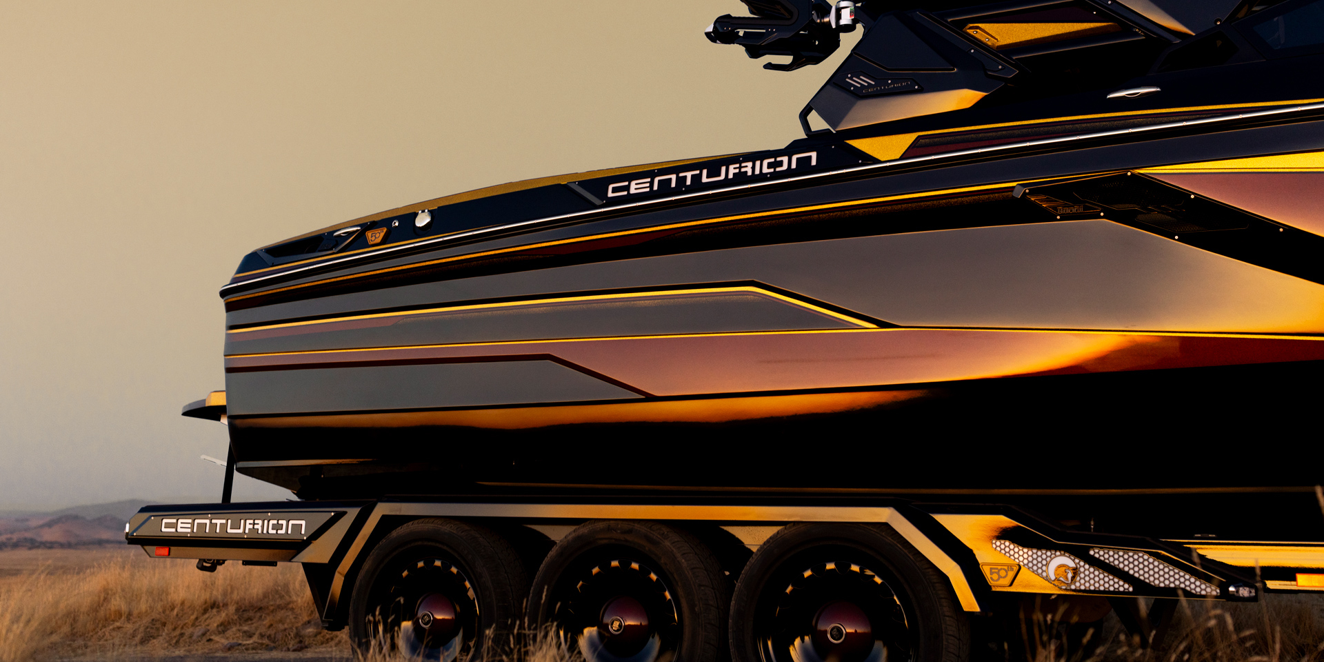 A close-up of a black and gold Centurion speedboat on a multi-axle trailer, parked on dry grass with a muted sky in the background.