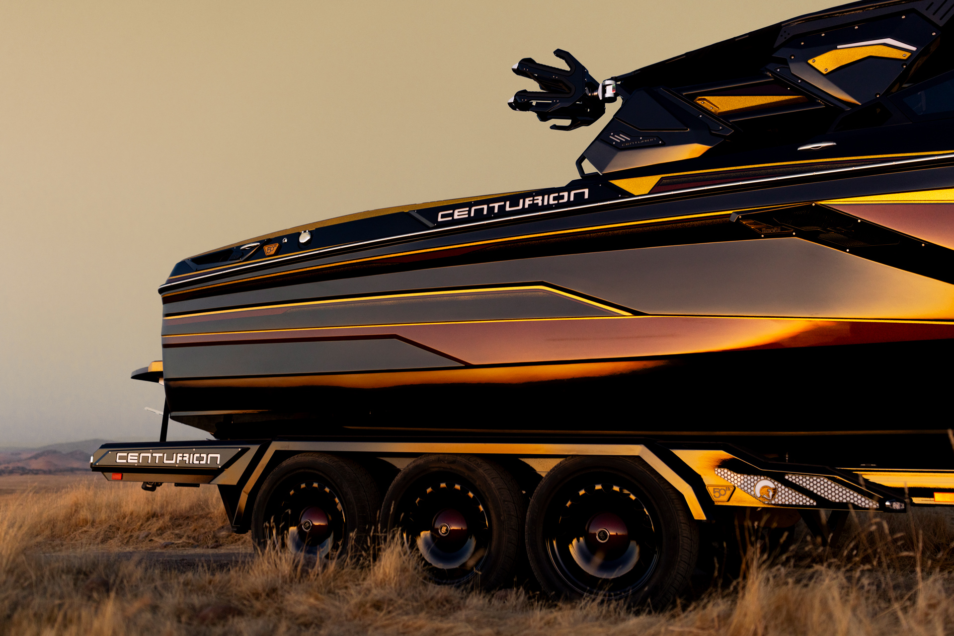 A close-up of a black and gold Centurion speedboat on a multi-axle trailer, parked on dry grass with a muted sky in the background.