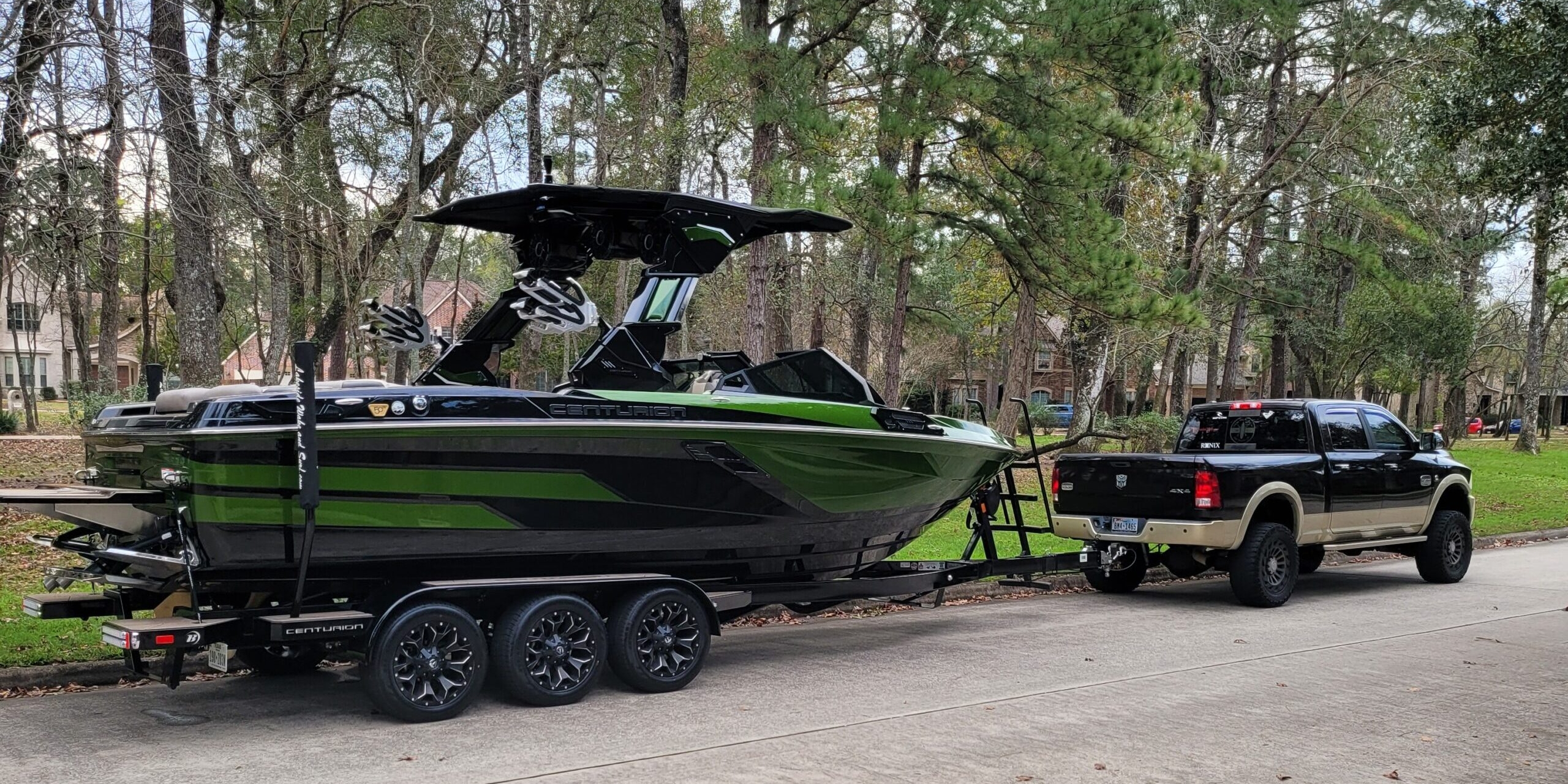 A black pickup truck is towing a large black and green speedboat on a trailer along a residential street lined with trees.
