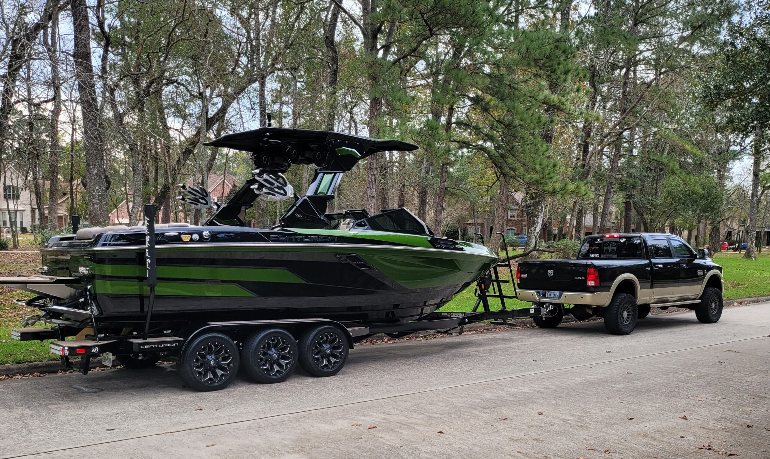 A black pickup truck is towing a large black and green speedboat on a trailer along a residential street lined with trees.