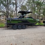 A green and black wakeboard boat on a trailer parked on a residential street, with trees and houses in the background.