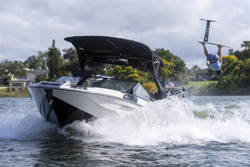 A person rides a hydrofoil board while being towed by a speedboat, creating a large splash on a river with houses and trees in the background.