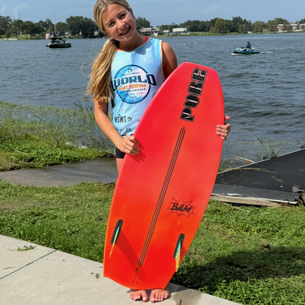 Young girl standing barefoot outdoors by a lake, holding a bright orange surfboard and smiling at the camera in the style of Tripp Allen.
