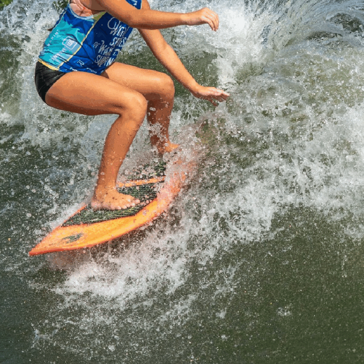 A young woman in a blue top and black shorts rides a wave on an orange surfboard, with trees and a cloudy sky in the background, capturing the adventurous spirit of Tripp Allen.