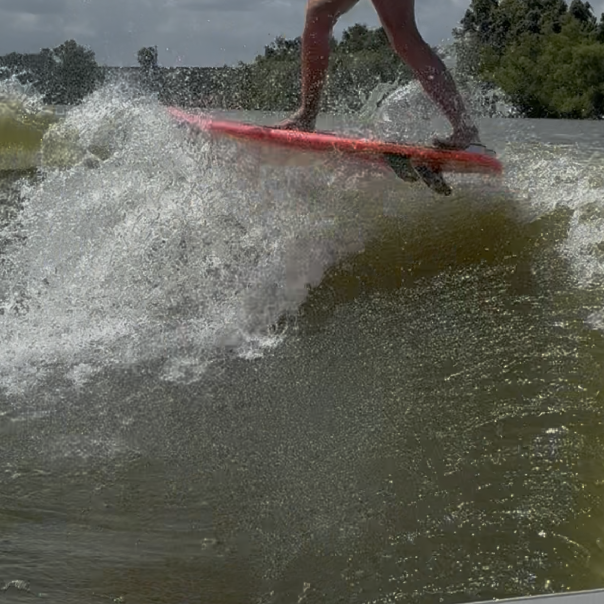 Tripp Allen, wearing a life jacket and shorts, is surfing on a wave—balancing on a red surfboard with water splashing around him, under a partly cloudy sky.