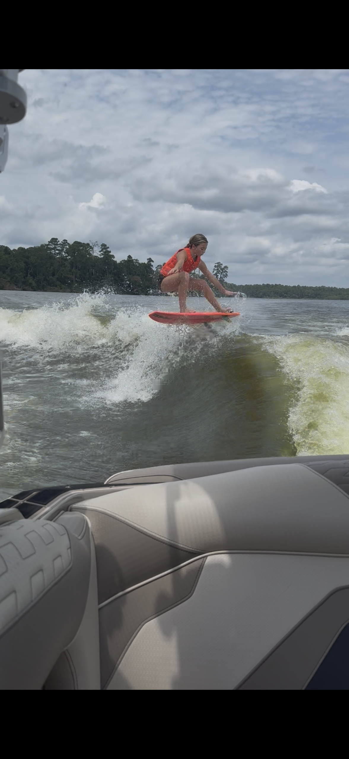 Tripp Allen, in a red life jacket, wakesurfs on a lake, riding a wave behind a boat beneath a cloudy sky.