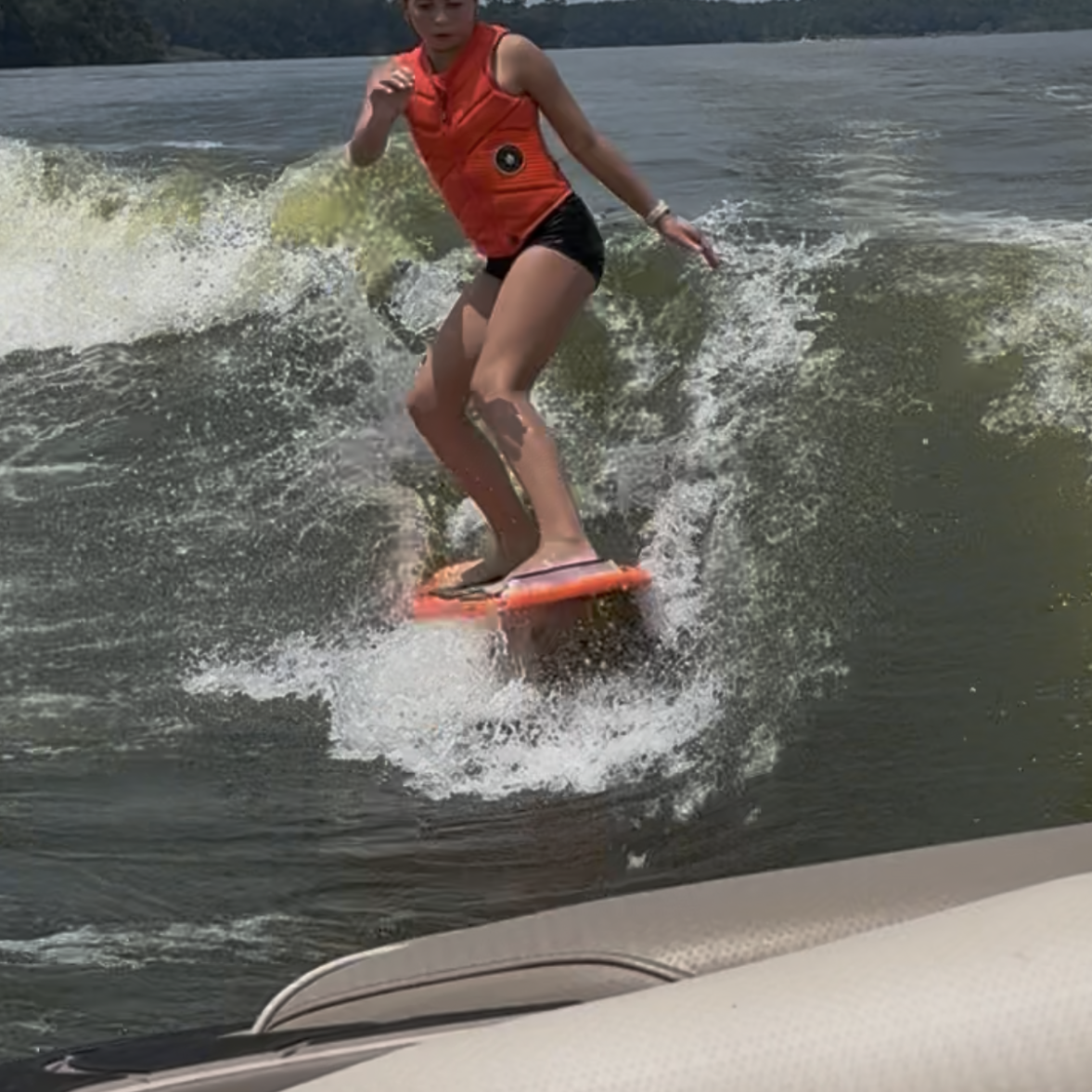 Tripp Allen, wearing an orange life jacket, is wake surfing on a lake behind a boat, with water splashing around and trees visible in the background.