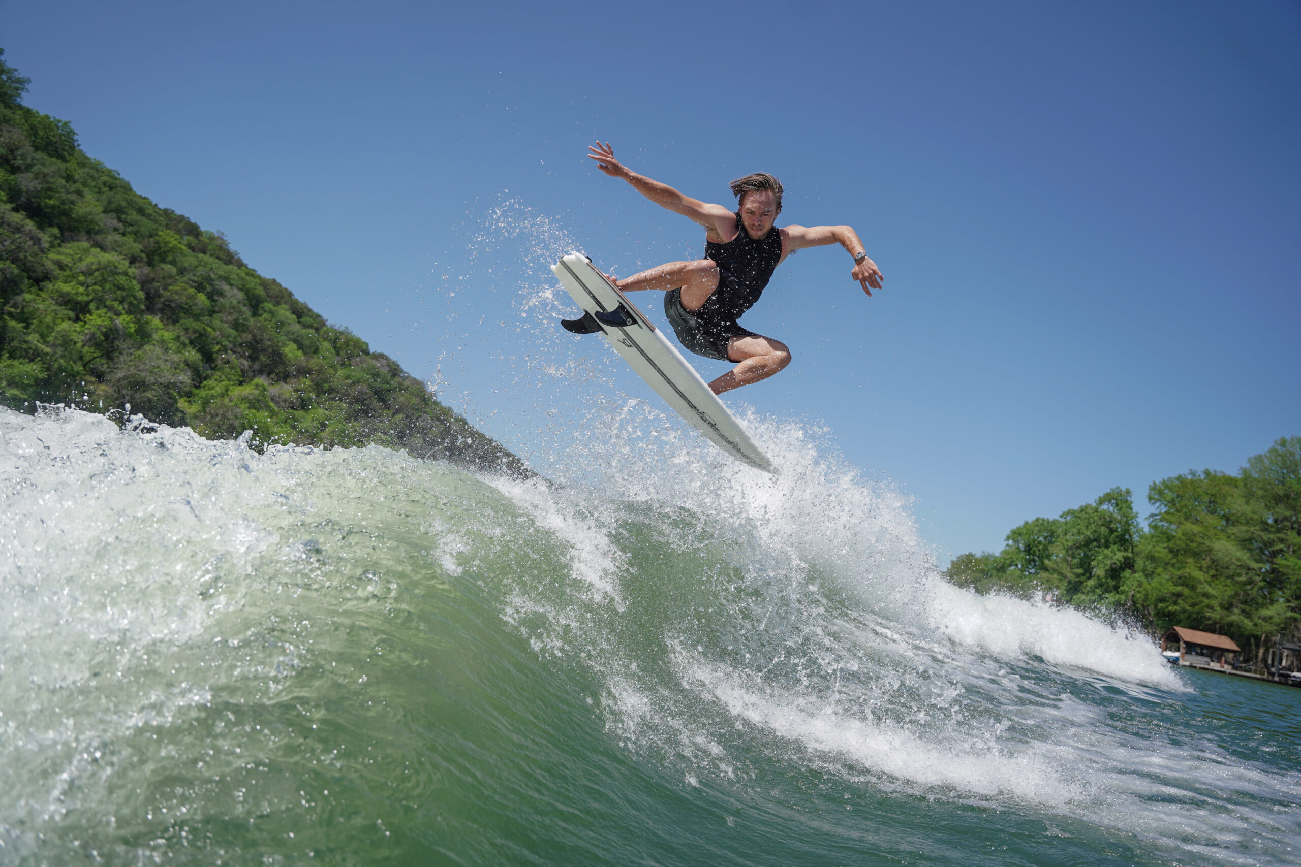 Trevor Miller, in a black wetsuit, jumps with a surfboard above a wave near a green, tree-lined shore under a clear blue sky.