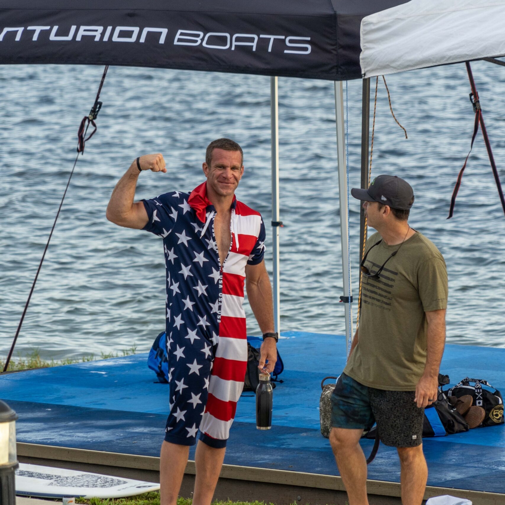 A man wearing an American flag outfit flexes his arm and smiles, standing barefoot near a tent by the water, while Clair Hurta in casual clothes stands beside him.