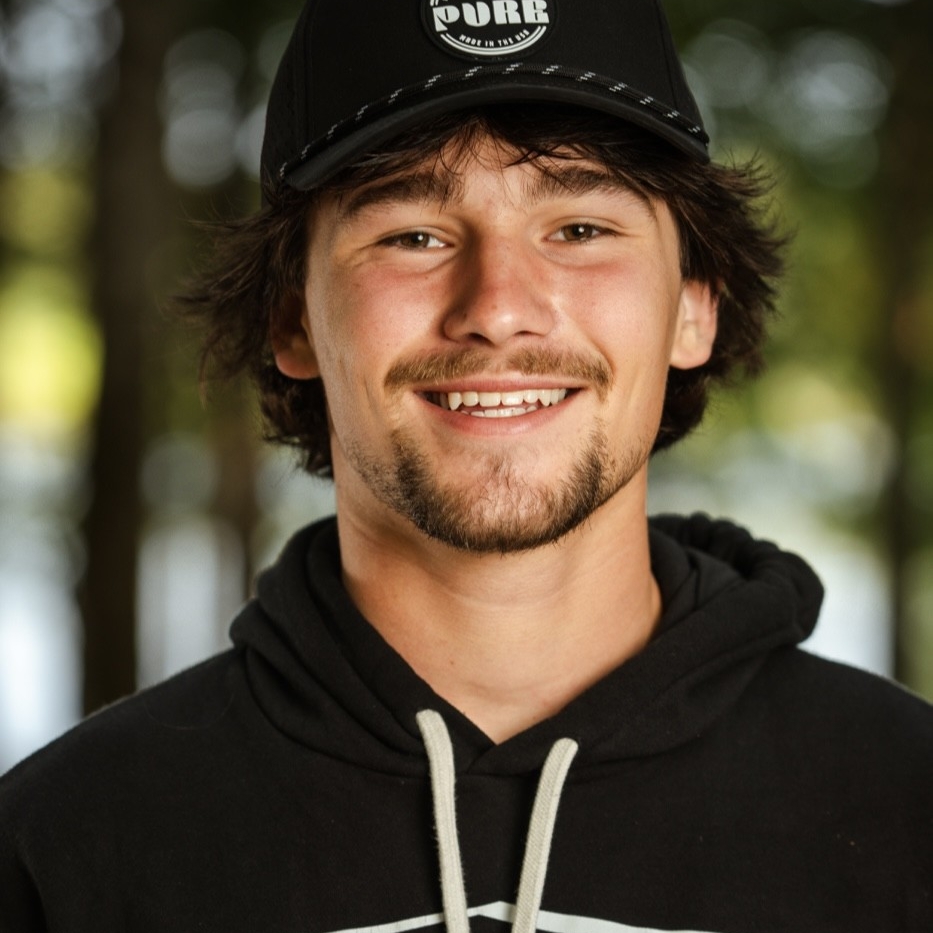 Jordan Koeninger, a young man with a short beard and mustache, wears a black hoodie and black cap, smiling in front of a blurred outdoor background with trees.