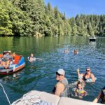 A group of people relax on a lake, some on a boat, some swimming, and some in a floating tube, surrounded by trees and clear blue water under a sunny sky.