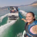 A woman in a life vest smiles and takes a selfie while wakeboarding, holding a rope behind a boat with several people on a lake surrounded by hills.