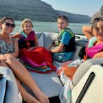Four people, including an adult woman and three children in life jackets, sit and smile on a boat in a canyon lake under sunny weather.
