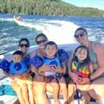 Three women and three young children wearing swimsuits and life jackets sit on a boat with a forested lake shore and people tubing in the background.