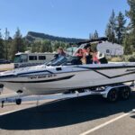 Three people sit in a white speedboat on a trailer in a parking lot, surrounded by trees and hills, with an RV visible in the background.