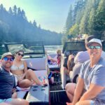 Four adults and a child relax on a motorboat on a sunny day, surrounded by water and forested hills.