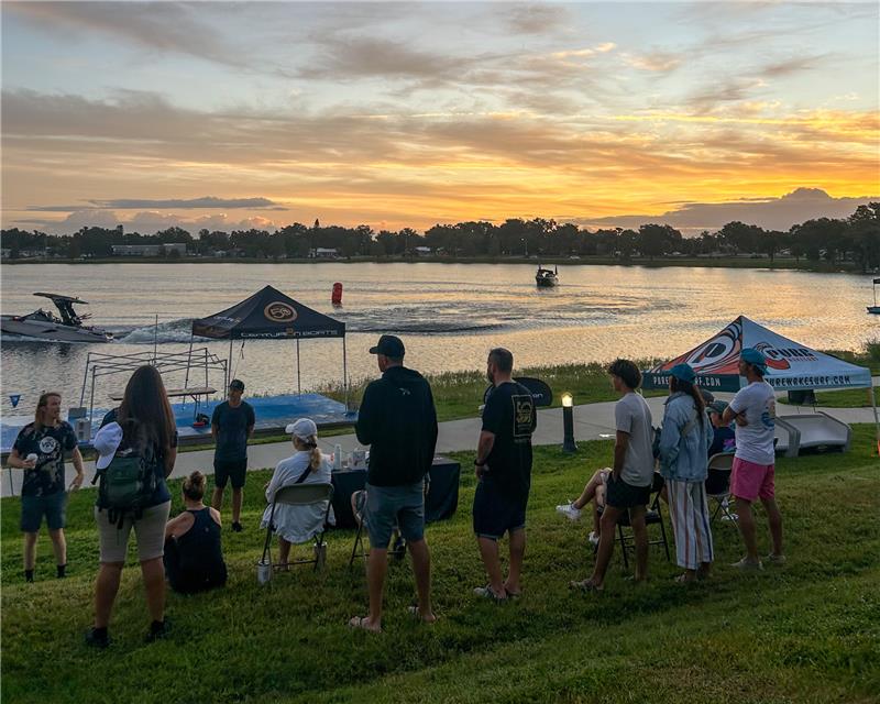 A group of people stand and sit near tents by a lakeside at sunset, with boats on the water and trees in the background.