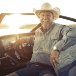 Smiling man in a cowboy hat sits at the helm of a modern boat at sunset, wearing a button-up shirt and shorts.