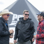 Four men stand outdoors talking and smiling near a large tent and a body of water, with one holding a can and another holding a bottle.