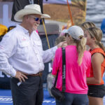 A man in a white shirt and cowboy hat talks to three young girls at an outdoor event near water.