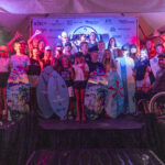 A large group of people, some holding surfboards, pose together in front of a sponsor backdrop under a tent at night; one person is in a wheelchair.