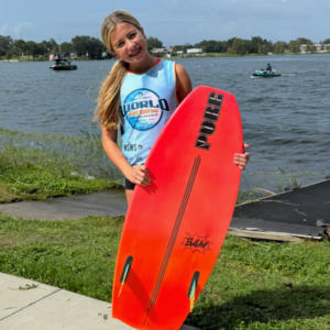 A girl stands on grass by a lake holding a bright orange wakesurf board, wearing a blue tank top with a “World Wake Surfing” logo.