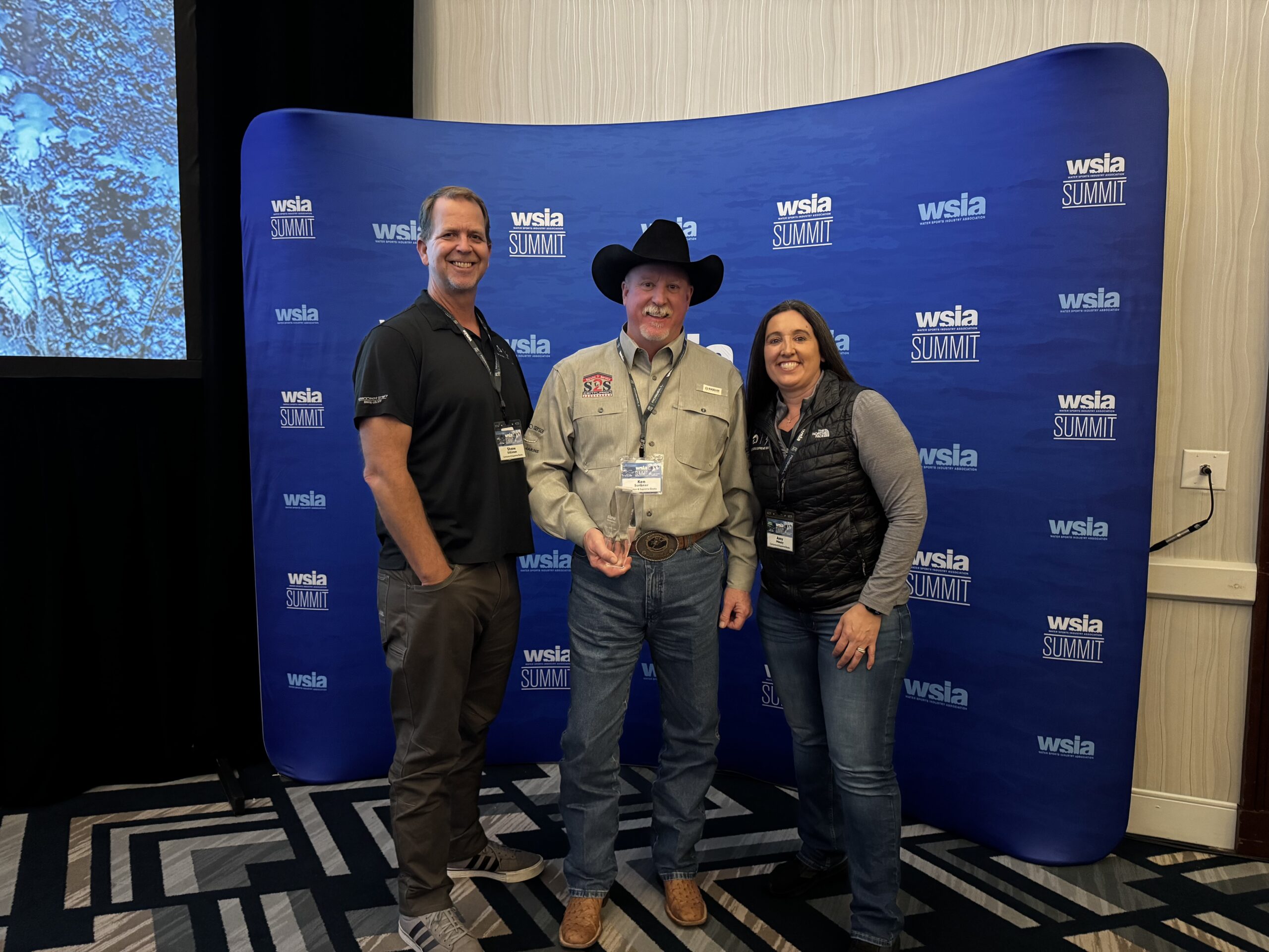 Three people pose together in front of a blue "wsia summit" backdrop; the man in the middle holds an award and wears a cowboy hat.