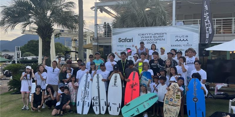 A group of people pose together outdoors in front of a building, holding surfboards at the Centurion Wake Surf Japan Open 2024 event.