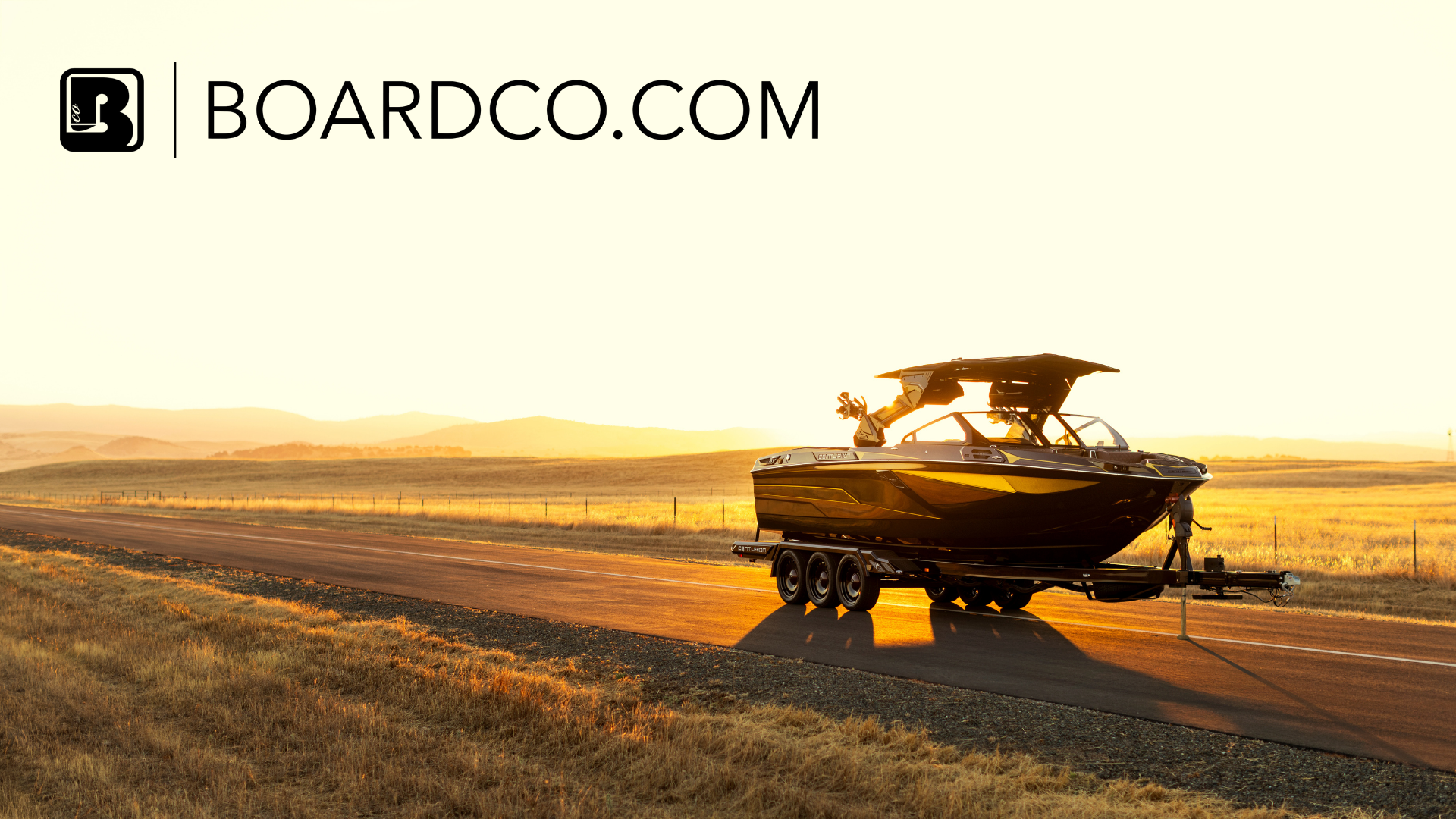 A black boat on a trailer is parked on a rural road at sunset, with fields and hills in the background. The text "BOARDCO.COM" is displayed at the top left.