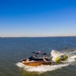 A person wake surfing behind a brown motorboat on a large, calm body of water under a clear blue sky.
